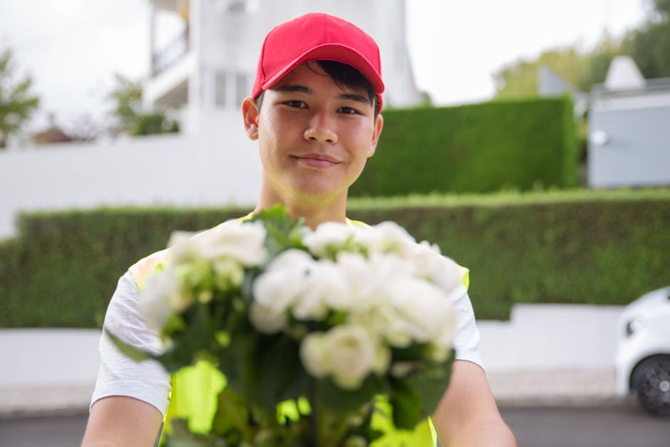 A young delivery person wearing a red cap and a neon yellow safety vest holds out a bouquet of fresh white flowers, including roses and peonies, wrapped in clear cellophane, with green foliage accents