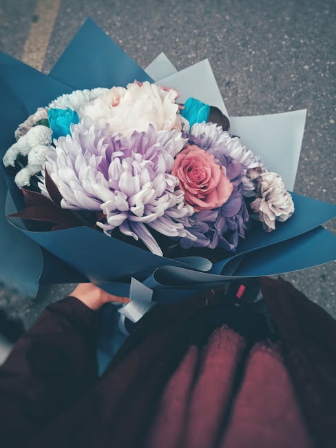 A hand holding a beautifully arranged floral bouquet featuring large, soft-petaled white chrysanthemums, complemented by pink roses and smaller purple and blue accent flowers. The bouquet is wrapped i