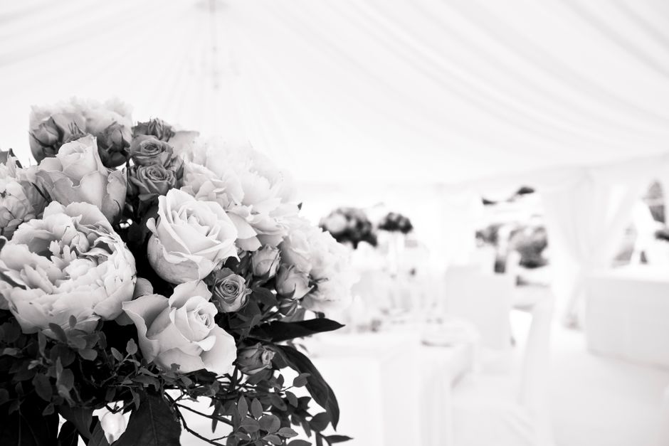 A close-up view of a floral arrangement featuring a lush bouquet of pale roses, peonies, and small filler blooms wrapped in white paper and green foliage, placed on a table inside a bright, elegant ma