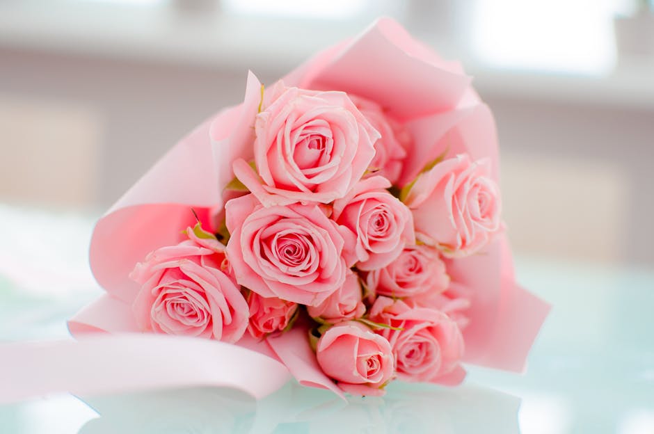 A bouquet of fresh pink roses arranged in a compact, rounded style, with their petals delicately layered and slightly curled at the edges. The roses are wrapped in soft pink tissue paper and secured w
