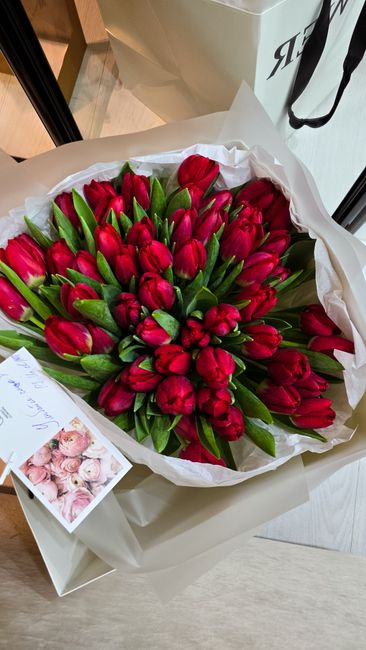 A bouquet of fresh red tulips with vibrant green leaves, arranged in a dense, rounded cluster. The tulips are wrapped in white tissue paper and placed in a white box, ready for flower delivery. The ar
